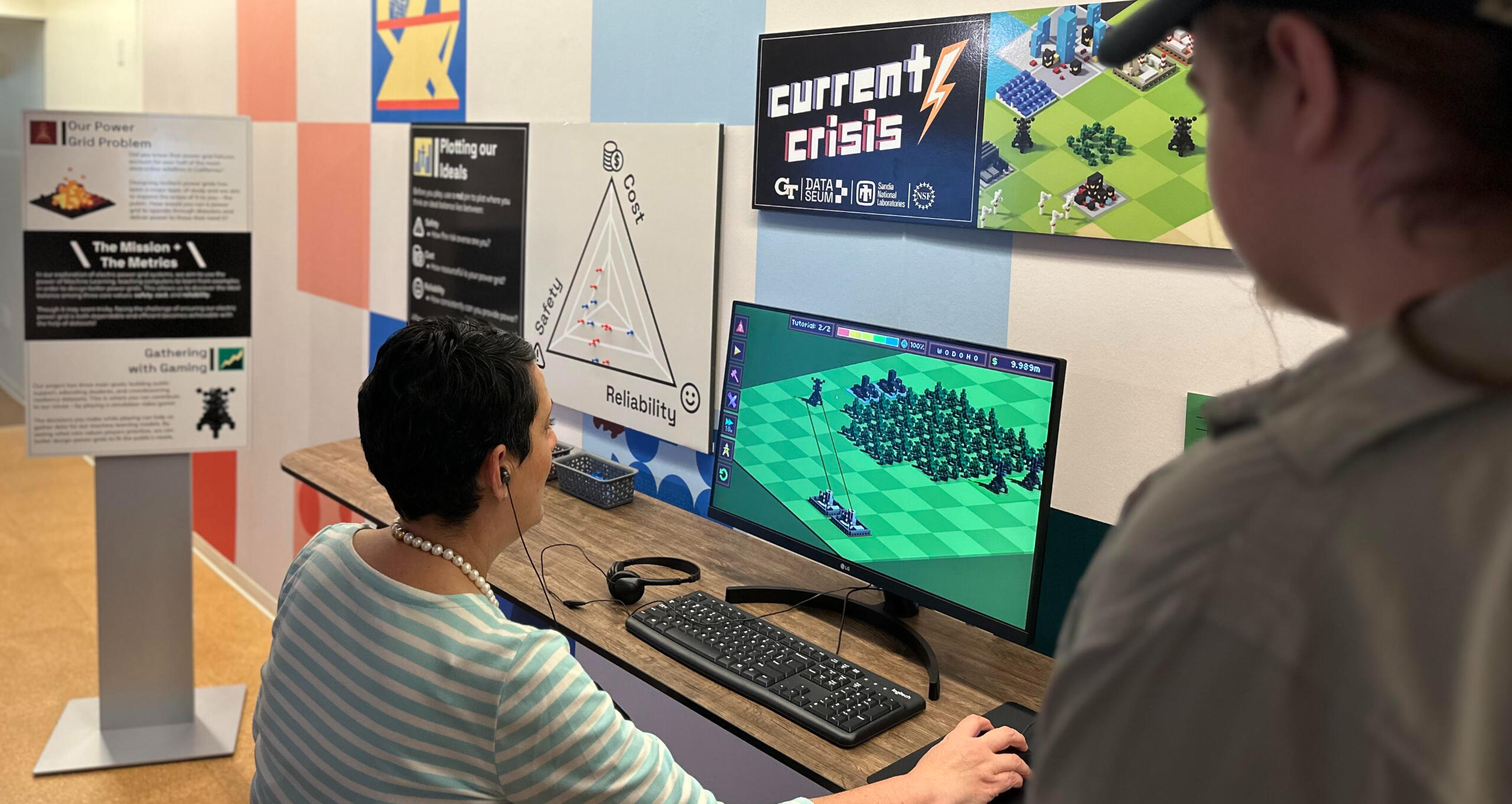 Person playing Current Crisis at a PC setup. 
                                        Various foam boards on display give background information on the game.